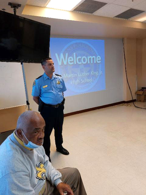 an officer from the New Orleans Police Department and a teacher in front of a project with the text: Welcome, Martin Luther King, Jr. High School
